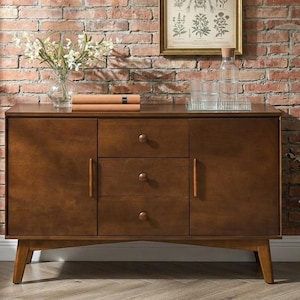 May include: A mid-century modern style wooden sideboard with three drawers and two cabinet doors. The sideboard is a warm brown color and has tapered legs. A vase of white flowers, books, and glassware are on top of the sideboard.