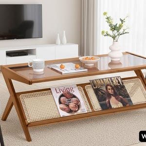 May include: A wooden coffee table with a clear glass top and a lower shelf with woven detailing. Magazines are displayed on the shelf. The table has a warm brown finish and is in a living room setting.