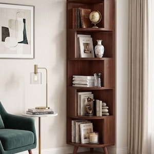 May include: A dark brown corner bookshelf with curved shelves. The shelves display books, a globe, a vase, and decorative items. A small side table with a lamp and a green armchair are in the foreground. The room has a neutral color palette.