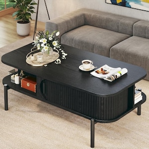 May include: A black wooden coffee table with a lower shelf and a cabinet door. The table has a dark finish and is furnished with a tray of flowers, a cup of coffee, and a magazine. The table is supported by black metal legs.