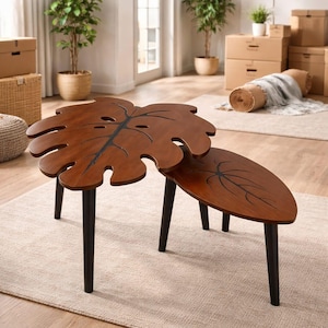 May include: Two-piece wooden leaf-shaped coffee table set. The tables have a dark brown finish with black accents and legs. The larger table is shaped like a monstera leaf, and the smaller table is oval.