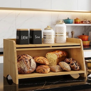 May include: A bamboo bread box with various types of bread and pastries. Two black canisters labeled "Coffee" and "Tea" sit on top, along with two decorative jars. The bread box is on a black countertop.