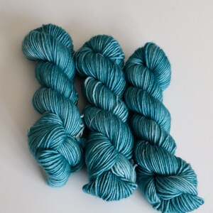 May include: Three skeins of teal yarn with white strands are displayed on a white surface. The yarn is tightly wound, showing a textured appearance. Suitable for knitting or crochet projects.