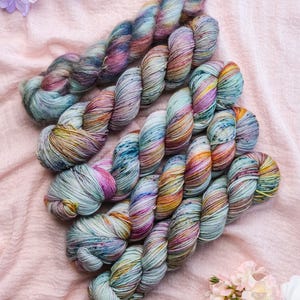 Puede incluir: Varios ovillos de hilo teñido a mano en una variedad de colores, incluyendo turquesa, rosa, amarillo y naranja. El hilo está dispuesto sobre una tela rosa con flores artificiales.