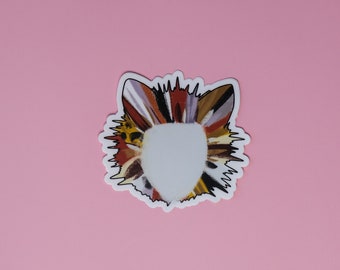 May include: A sticker featuring a stylized cat face with a white center and a colorful, textured design in shades of brown, orange, and purple. The sticker has a black outline and is set against a pink background.