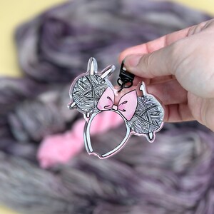 May include: A clear plastic charm shaped like Mickey Mouse ears with a pink bow. The ears are filled with a black and white yarn design.