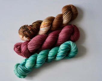 May include: Four skeins of yarn in different colours and patterns. The yarn is arranged in a pile, with the ends of the skeins overlapping. The colours are brown, pink, green, and a multi-coloured brown and white.
