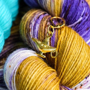 Puede incluir: Un amuleto de lámpara de genio dorado unido a un ovillo de hilo morado, blanco y dorado. El hilo está enrollado en forma de bola.