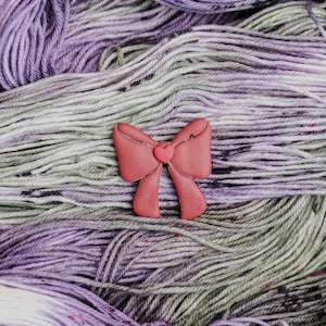 May include: A close-up shot of a skein of yarn in shades of purple, gray, and white. A small, coral-colored bow-shaped embellishment with a heart in the center rests on top of the yarn. The yarn appears soft and textured.