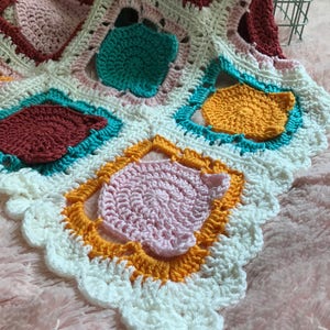 May include: A crocheted blanket with a white background and colorful squares in shades of pink, yellow, orange, teal, and burgundy. The blanket has a scalloped edge.
