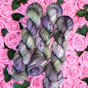 Può includere: Matasse di filato tinto a mano nei toni del viola, verde e grigio, disposte su un letto di rose rosa. Il filato ha un aspetto maculato e variegato, con sottili variazioni di colore. Lo sfondo è una fitta disposizione di rose rosa.