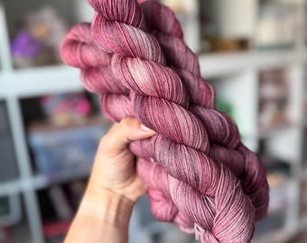May include: A skein of hand-dyed yarn in shades of pink, burgundy, and cream. The yarn is held up, showcasing its soft texture and colour variations. Ideal for knitting or crochet projects.