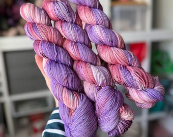 May include: Three skeins of yarn in shades of pink, purple, and white. The yarn is hand-dyed and has a speckled effect.