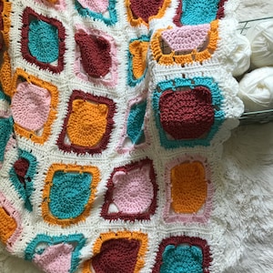 May include: A white crocheted blanket with a repeating pattern of colorful squares. Each square features a different color and a bear face design. The colors include pink, teal, orange, and brown.