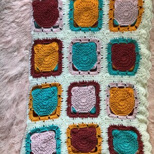 May include: A crocheted baby blanket with a white background and colorful squares. The squares are in shades of pink, teal, brown, and yellow.