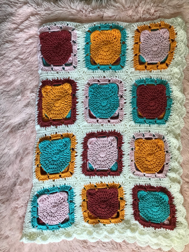 May include: A crocheted baby blanket with a white background and colorful squares. The squares are in shades of pink, teal, brown, and yellow.