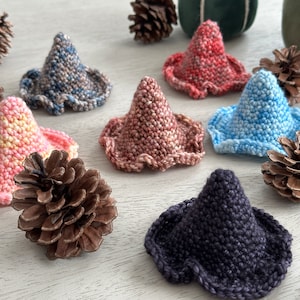 May include: Six crocheted witch hats in various colors, including pink, blue, brown, and gray, are arranged on a light wood surface.  The hats are all cone-shaped with a ruffled edge.  There are also pine cones scattered around the hats.