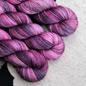 May include: Close-up of several skeins of hand-dyed yarn in shades of pink and purple. The yarn is arranged diagonally against a white wooden surface. A black fabric with a white pattern is visible in the background.