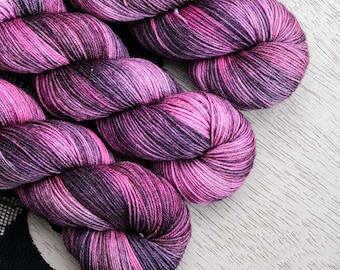 May include: Close-up of several skeins of hand-dyed yarn in shades of pink and purple. The yarn is arranged diagonally against a white wooden surface. A black fabric with a white pattern is visible in the background.