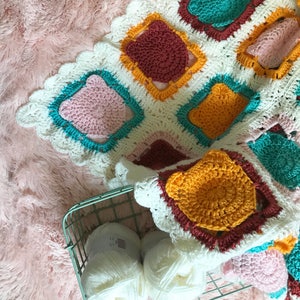 May include: A white crocheted blanket with colorful squares in shades of pink, teal, yellow, and brown. The blanket is folded and placed on a pink fuzzy surface. Two balls of white yarn are in a wire basket.