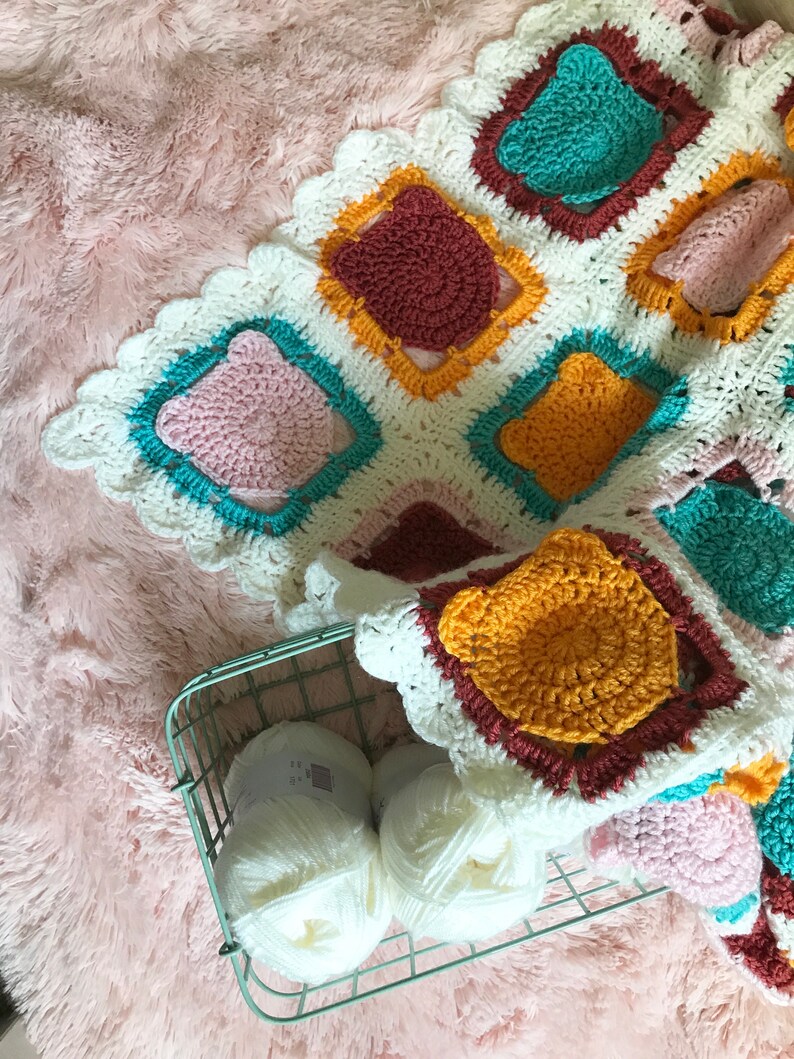 May include: A white crocheted blanket with colorful squares in shades of pink, teal, yellow, and brown. The blanket is folded and placed on a pink fuzzy surface. Two balls of white yarn are in a wire basket.