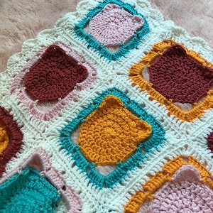 May include: A crocheted blanket with a white background and colorful squares. The squares are in shades of pink, teal, orange, and brown.