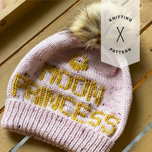 May include: Pink knitted hat with a yellow "Mochi Princess" inscription and a fluffy white pom pom on top. The hat is on a wooden surface.