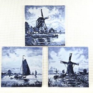 May include: Three square ceramic tiles with blue and white Dutch landscape scenes. Each tile depicts a windmill, a sailboat, or a waterside village. The tiles are arranged on a white textured surface.