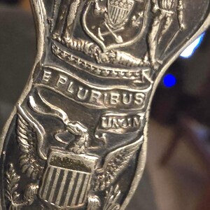 May include: Close-up of a silver-colored metal object with intricate embossed details. The design includes the words "FORWARD", "E PLURIBUS UNUM", and "NATIONAL UNION", along with images of figures and an eagle.