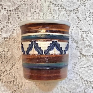 May include: A handcrafted ceramic mug with a brown, blue, and white geometric design. The mug features a cylindrical shape with a white rim and a glossy finish. The design includes horizontal bands and a repeating pattern.