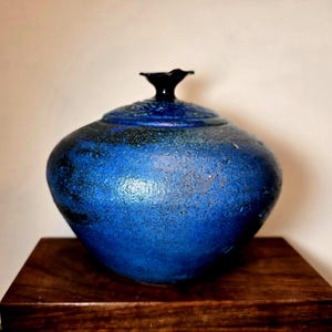 May include: A handcrafted, blue ceramic jar with a textured surface and a black, flower-shaped lid. The jar has a rounded body and sits on a dark wooden surface. The color transitions from a deep blue to a lighter shade, creating a unique aesthetic.