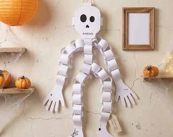 Könnte beinhalten: Eine Skelett-Dekoration aus weißen Papierketten mit einem Totenkopf, schwarzen Augen und einem genähten Mund. Die Gliedmaßen bestehen aus Papierringen, ergänzt durch Hand- und Fußausschnitte. Ein Fuß zeigt ein Spinnennetz, der andere eine Spinne.