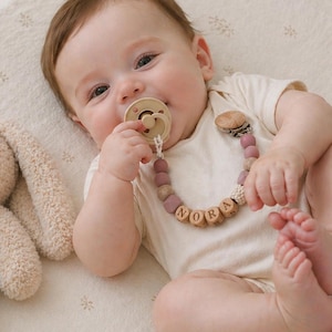 May include: A baby wearing a white onesie and a pacifier. The pacifier is attached to a beaded chain with the name "NORA" spelled out in wooden letters. A stuffed bunny is next to the baby.