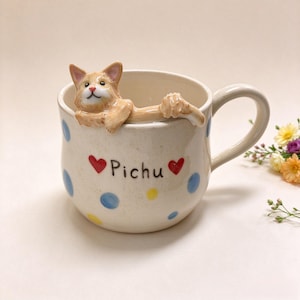 Puede incluir: Una taza de cerámica color crema con una figura tridimensional de un gato jengibre descansando en el borde. La taza está decorada con lunares azules y amarillos, dos corazones rojos y el nombre Pichu impreso en letras negras al frente.