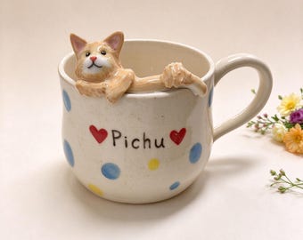 May include: A cream-colored ceramic mug featuring a three-dimensional ginger cat figurine resting on the rim. The mug is decorated with blue and yellow polka dots, two red hearts, and the name Pichu printed in black lettering on the front.