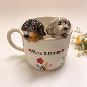 May include: A cream-colored ceramic mug featuring two sculpted dog figurines peering over the rim. One dog is black and tan, the other is light brown. The mug is decorated with small colorful flowers and the text Max & Daisy with red hearts.