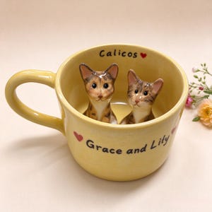 Puede incluir: Una taza de cerámica amarilla con dos cabezas de gato esculpidas en su interior. El exterior tiene la inscripción "Grace and Lity" junto a pequeños corazones rojos, mientras que el borde interior muestra la palabra "Calicos" con un corazón.