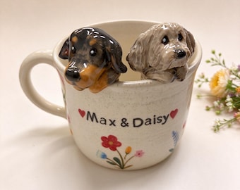 May include: A cream-colored ceramic mug featuring two sculpted dog figurines peering over the rim. One dog is black and tan, the other is light brown. The mug is decorated with small colorful flowers and the text Max & Daisy with red hearts.
