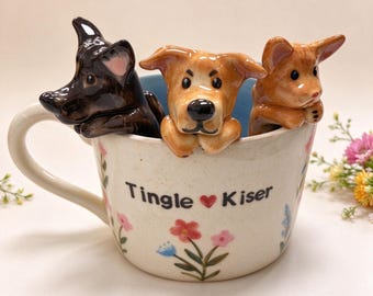 May include: A cream-colored ceramic mug featuring a floral pattern and the text Tingle Kiser with a red heart. Three sculpted dog figurines in black, tan, and light brown are perched on the rim, peering over the edge of the mug.