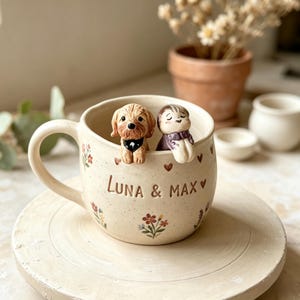 May include: A cream-colored ceramic mug with a speckled finish, featuring the text "Luna & Max" in brown letters. The mug is decorated with small painted flowers and hearts, and has two sculpted figures of a dog and a person resting on the rim.