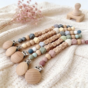 Puede incluir: Cuatro clips de chupete personalizados para bebés con cuentas de madera y detalles de silicona. Cada clip presenta un nombre diferente escrito con letras de madera. Los clips tienen una variedad de colores, incluyendo gris, beige y rosa.