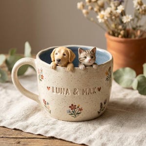 Könnte beinhalten: Ein cremefarbener Keramikbecher mit gesprenkelter Oberfläche, auf dessen Rand ein geformter Golden Retriever Welpe und ein getigertes Kätzchen sitzen. Der Becher ist mit kleinen Blumenmustern und dem Text LUNA & MAX sowie zwei kleinen roten Herzen verziert.