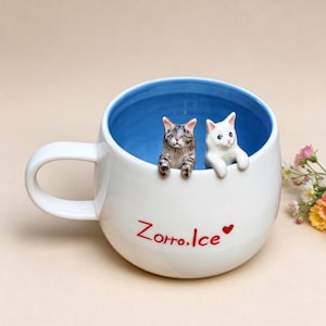 May include: A white ceramic mug with a bright blue interior. Two small, sculpted cat figures, one brown tabby and one white, are perched on the inner rim. The exterior features the red text "Zorro, Ice" followed by a small red heart symbol.