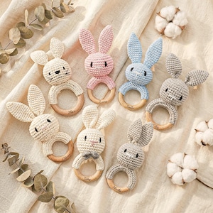 May include: A selection of handmade crochet bunny baby rattles in cream, pink, blue, and grey. Each rattle has a wooden ring and is personalised with a name. The bunnies have long ears and embroidered facial features.