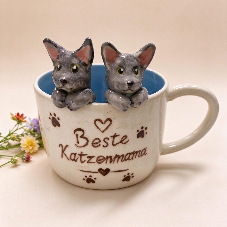 May include: A ceramic mug with a light blue interior and cream-colored exterior. Two sculpted gray cat figures peek over the rim. The front features brown text reading "Beste Katzenmama" with heart and paw print accents.