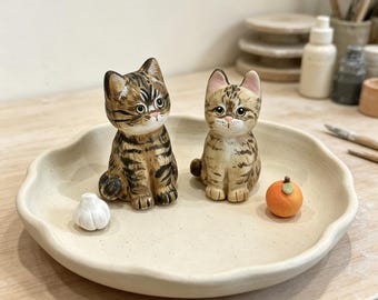 May include: Two ceramic cat figurines, one dark brown tabby and one light tan tabby, sit on a round, off-white ceramic plate with a wavy edge. A small white garlic bulb and a miniature orange fruit are placed on the plate next to the cats.