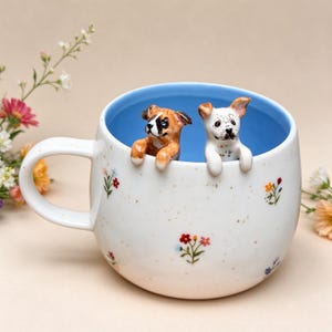 May include: A white ceramic mug with a light blue interior, featuring small colorful floral patterns and brown speckles on the exterior. Two small, sculpted dog figurines, one brown and one white with spots, peek over the rim.