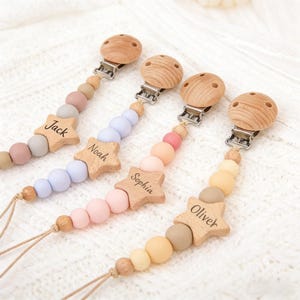 May include: Four baby pacifier clips with wooden star-shaped name tags. Each clip has a wooden clip and a string of colorful beads in shades of pink, blue, beige, and yellow. The names "Jack", "Noah", "Sophia", and "Oliver" are engraved on the stars.