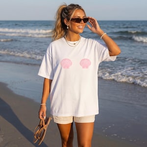 Puede incluir: Camiseta blanca con dos diseños de conchas marinas rosas. La camiseta es de estilo casual y oversize. La imagen muestra a una persona usando la camiseta en una playa, sugiriendo un tema de verano o playa.