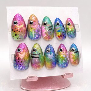 May include: A set of ten rainbow-colored, glittery, oval-shaped press-on nails. Each nail features a fish design with a smiling mouth, an eye, and various patterns in pink, black, and blue. The nails are displayed on a white card.